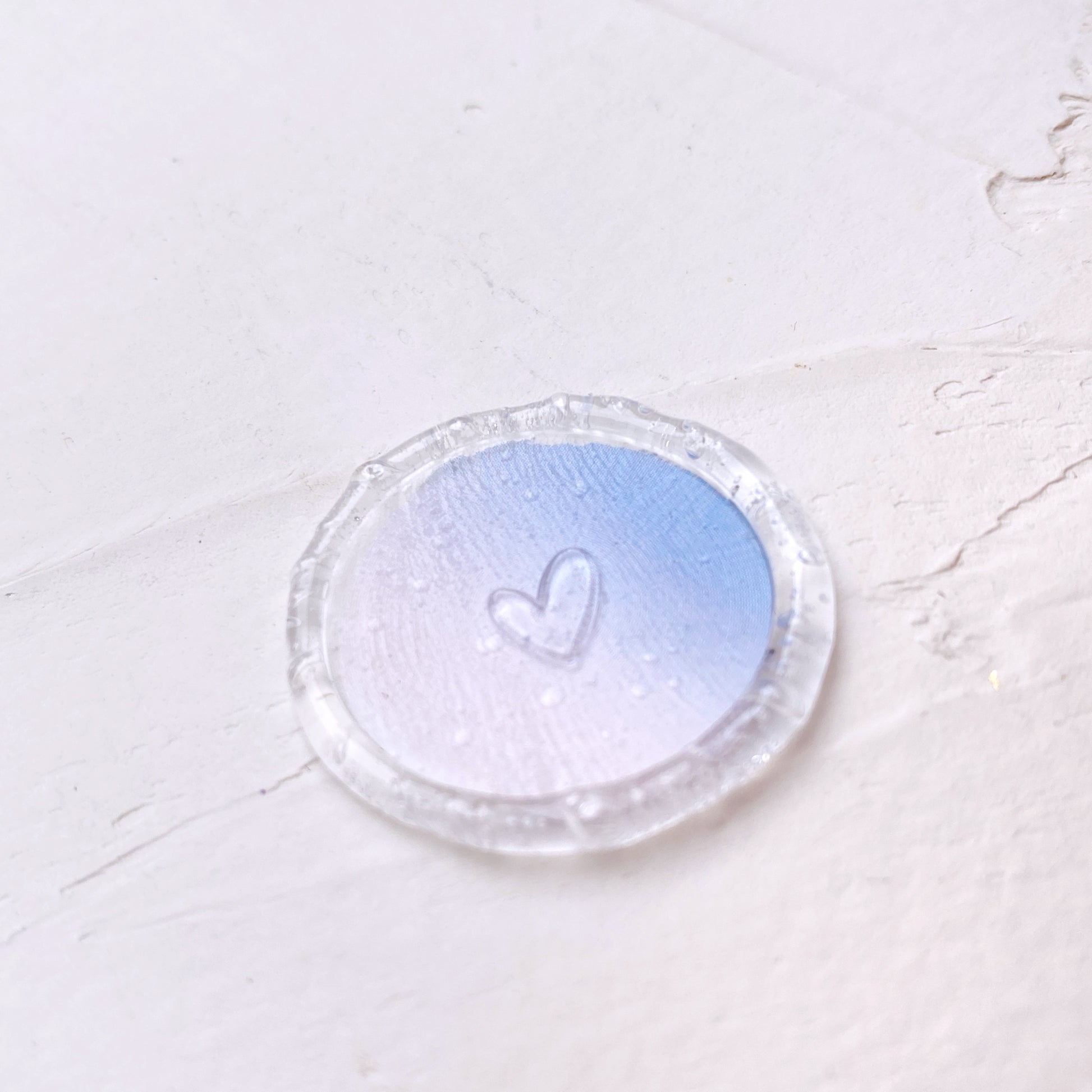 Průhledná pečeť se srdíčkem a modro-růžovým výsekem na svatební oznámení na bílém podkladu detail zeshora
