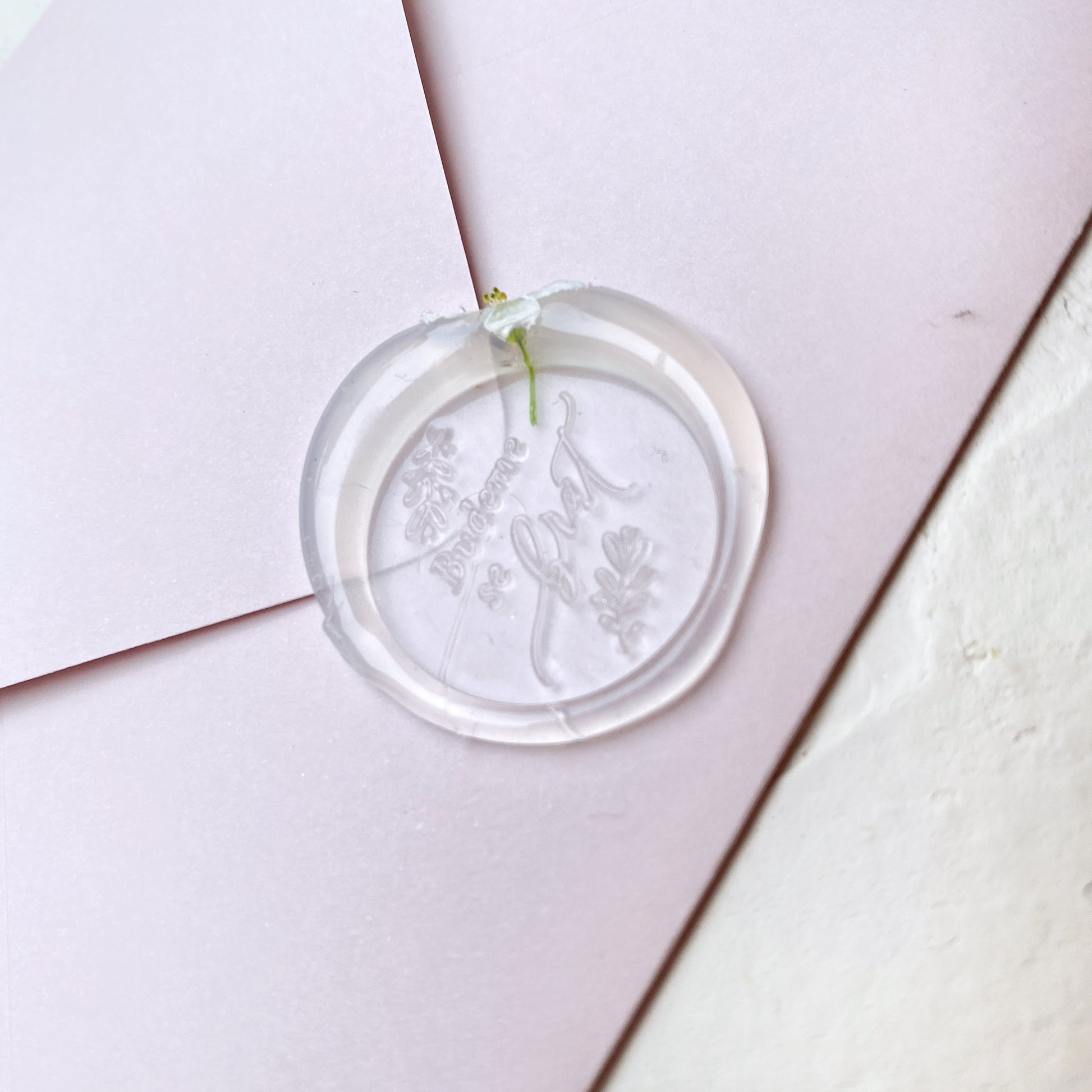 Průhledná svatební pečeť s květinou a textem "budeme se brát" na růžové obálce a bílém podkladu detail z profilu