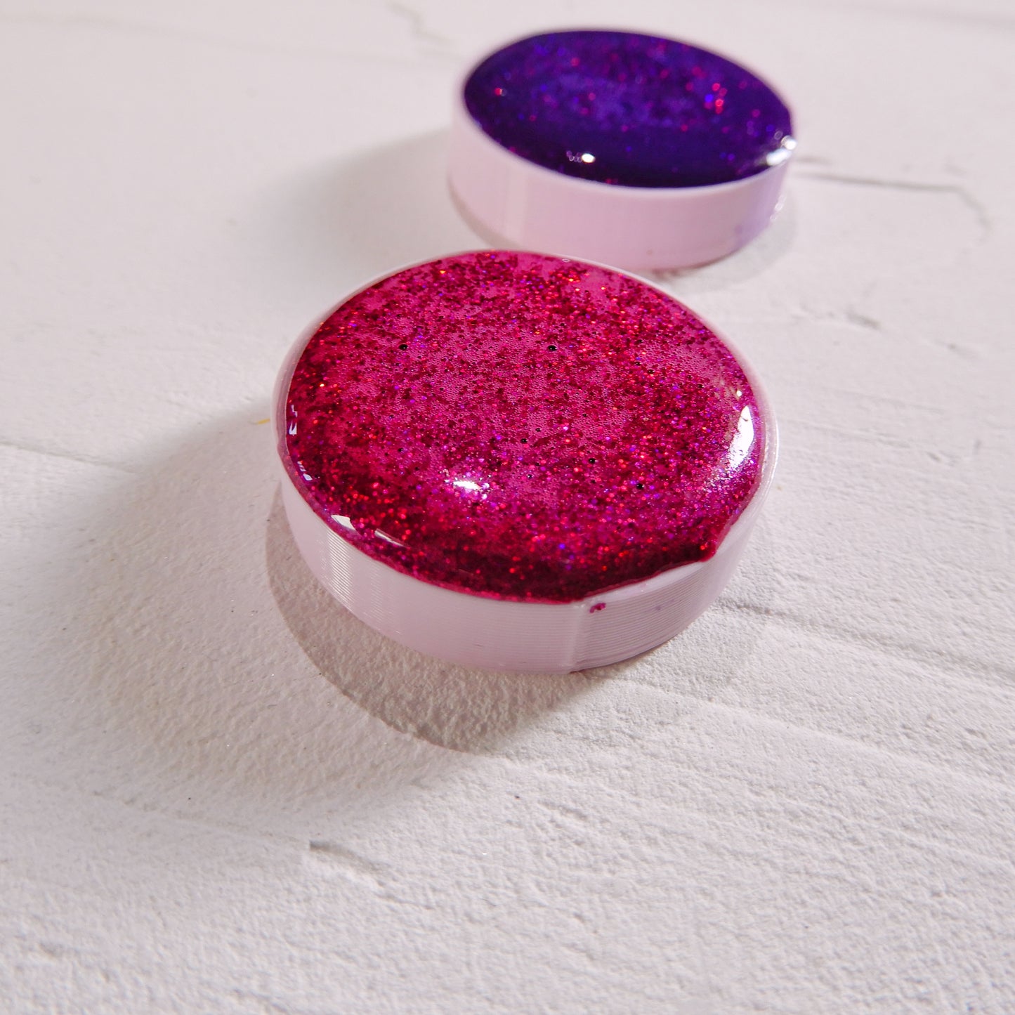 Ručně vyráběné akvarelové barvy se třpytkami v růžové a holografické fialové variantě na bílém podkladu zeshora